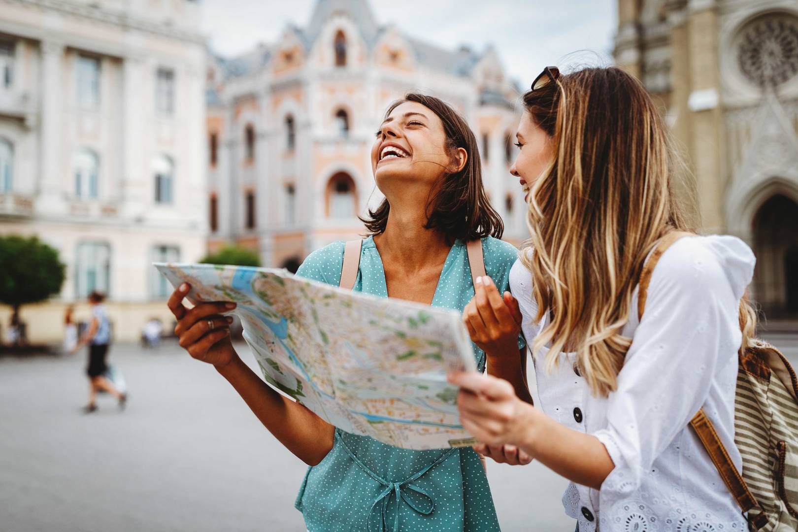 Two people stand in a city square holding and looking at a paper map, with historic buildings in the background. One person wears a teal dress and the other wears a white top with a backpack; both are positioned in the foreground while pedestrians and ornate architecture appear in soft focus behind them.