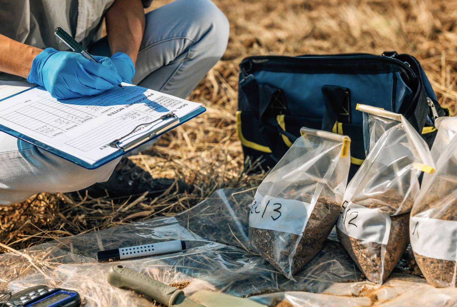 Gloved hand writing on a clipboard beside numbered plastic bags of soil samples and sampling equipment on dry ground