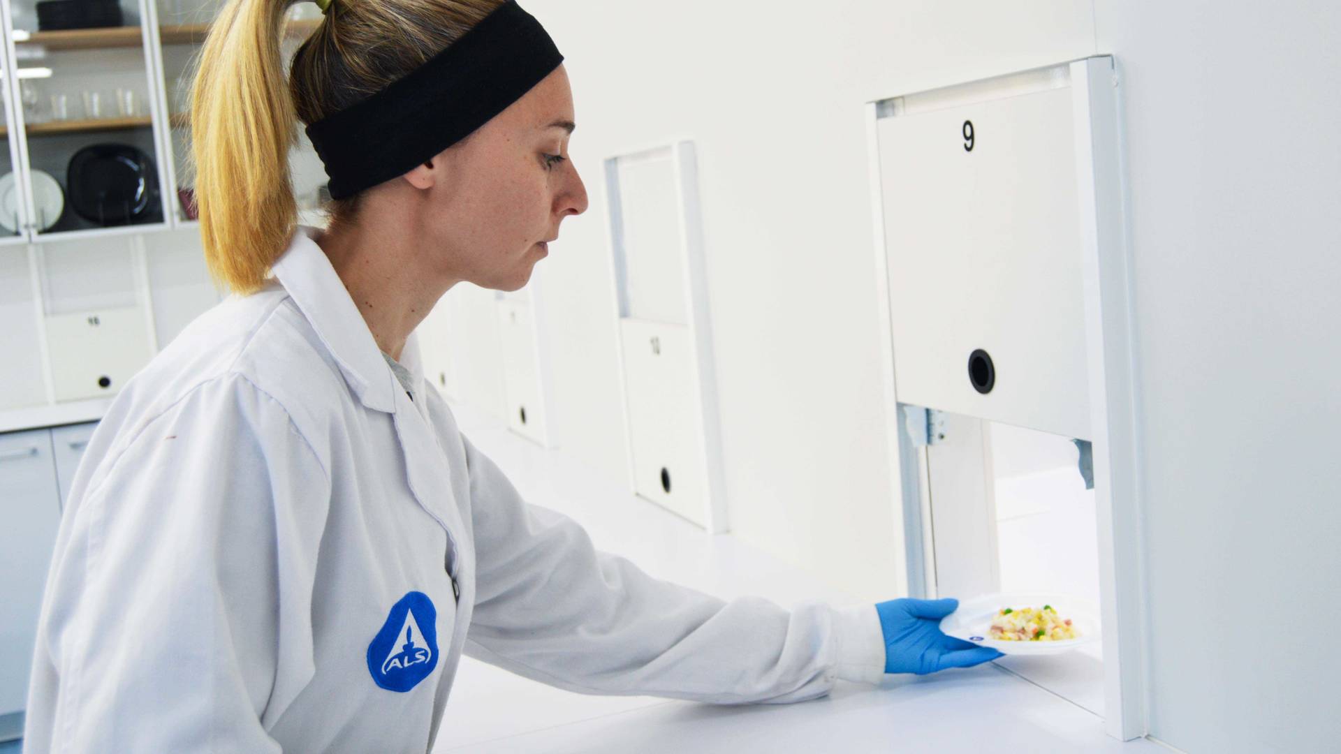 Osoba w białym fartuchu laboratoryjnym z logo ALS i w niebieskich rękawicach ochronnych umieszcza mały talerz z próbką jedzenia w numerowanym okienku przekazowym w sterylnym pomieszczeniu. W tle widoczne są białe ściany i kilka podobnych przegródek.
