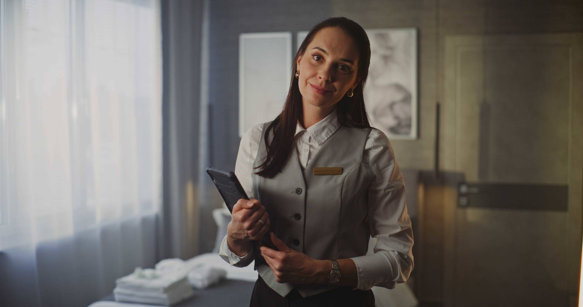 Person wearing a uniform and name badge, holding a tablet in a softly lit room with folded towels and framed wall art in the background.