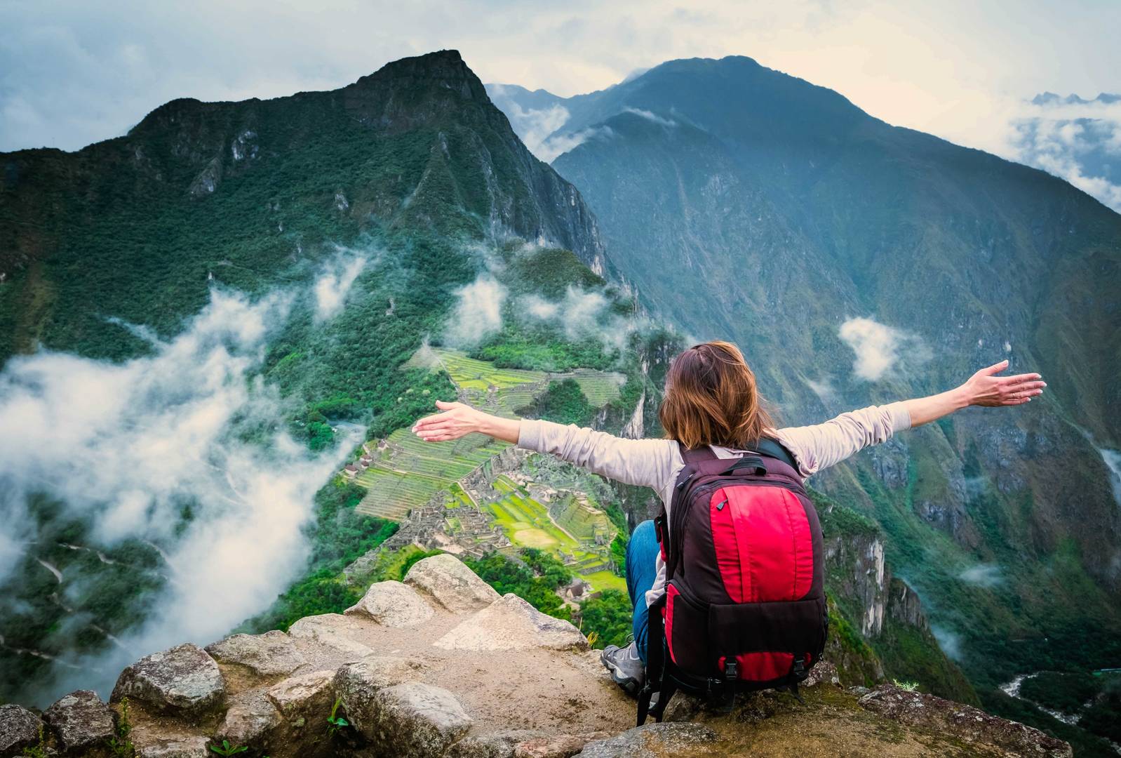 A person with a red backpack sits on a rocky ledge with arms outstretched, overlooking a lush green mountain valley with steep peaks, terraced fields, and scattered ruins partially covered by drifting clouds.