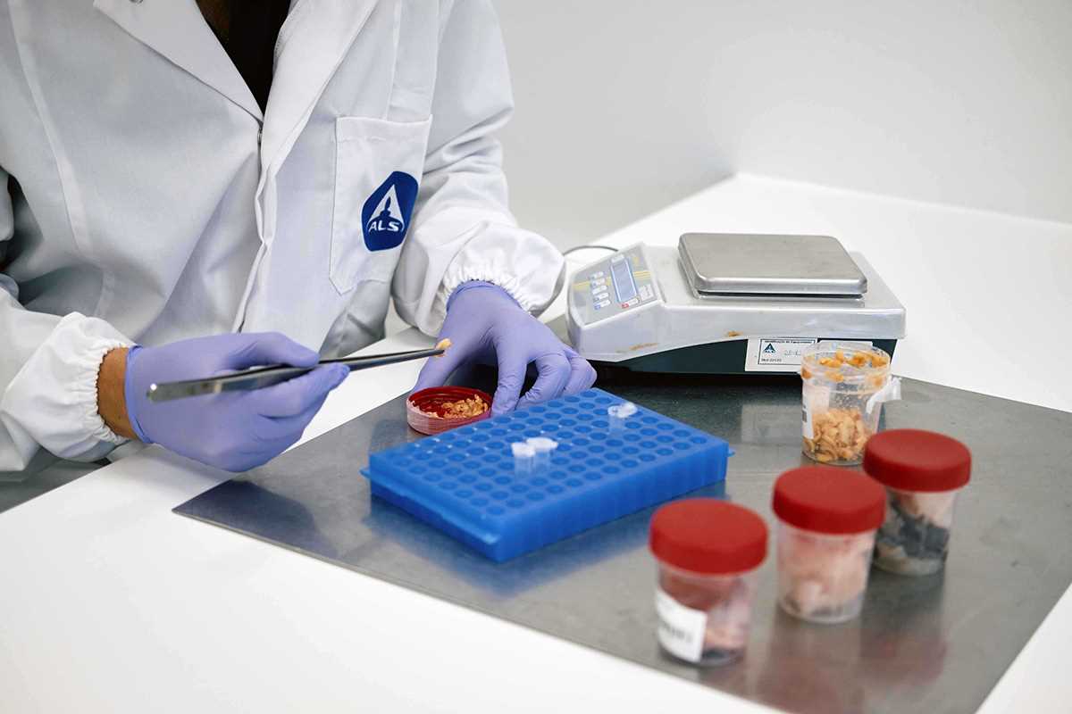 Técnico de laboratório com bata ALS e luvas roxas a utilizar pinça para manipular amostras junto a balança eletrónica.