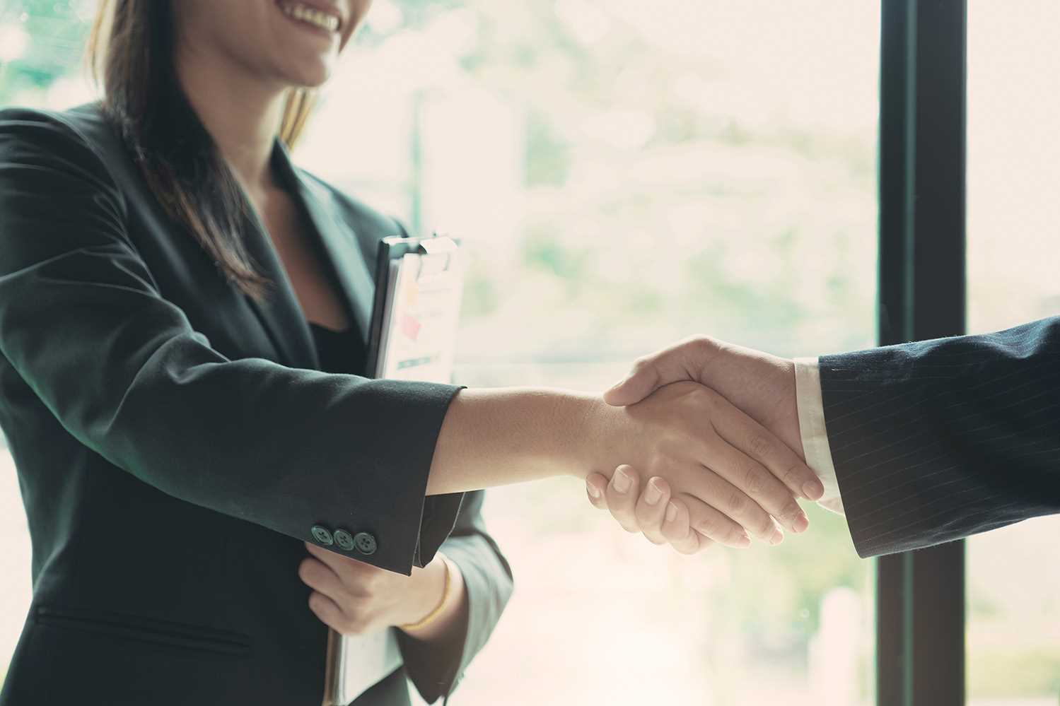 Dois profissionais em fato escuro a apertar as mãos, um deles com uma prancheta, num ambiente interior com luz natural.