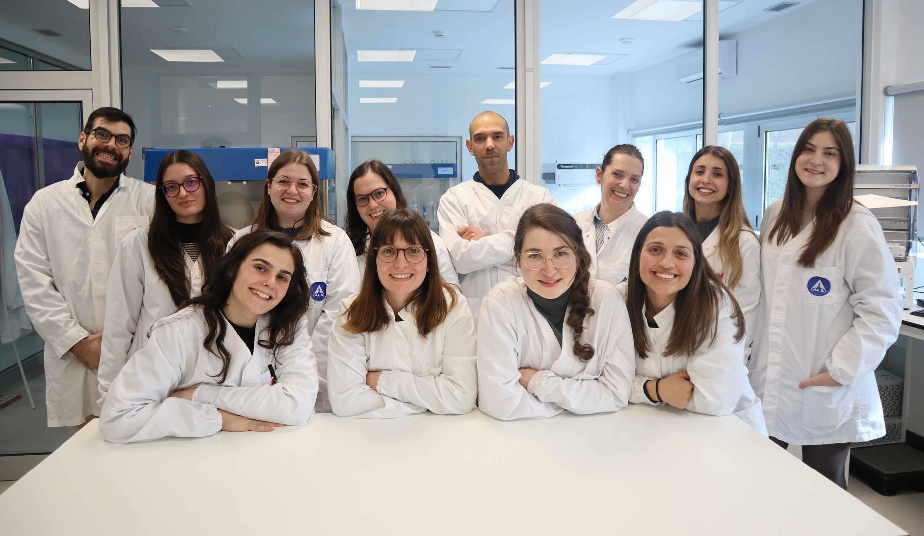 Grupo de pessoas com batas brancas sentadas e em pé à volta de uma mesa num ambiente de laboratório. 