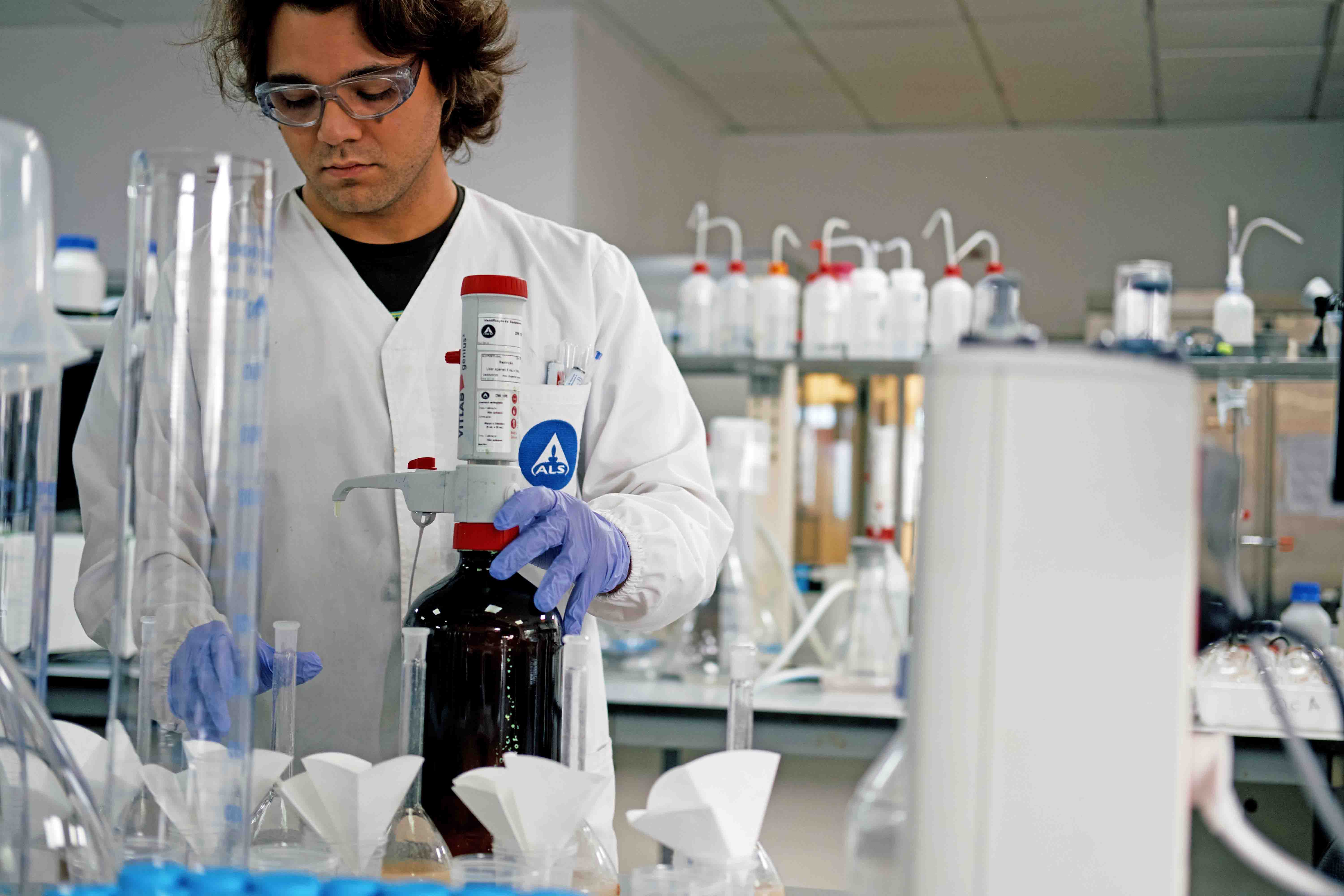 Pessoa com luvas e bata branca a utilizar frasco castanho com doseador num laboratório.