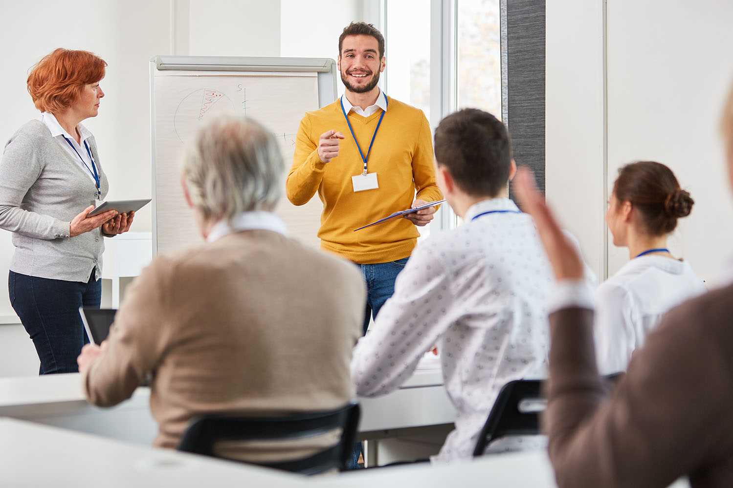 Grupo de pessoas a assistir a uma apresentação numa sala de formação. Uma pessoa está de pé à frente com um caderno, enquanto outra segura um tablet. O público está sentado e atento.