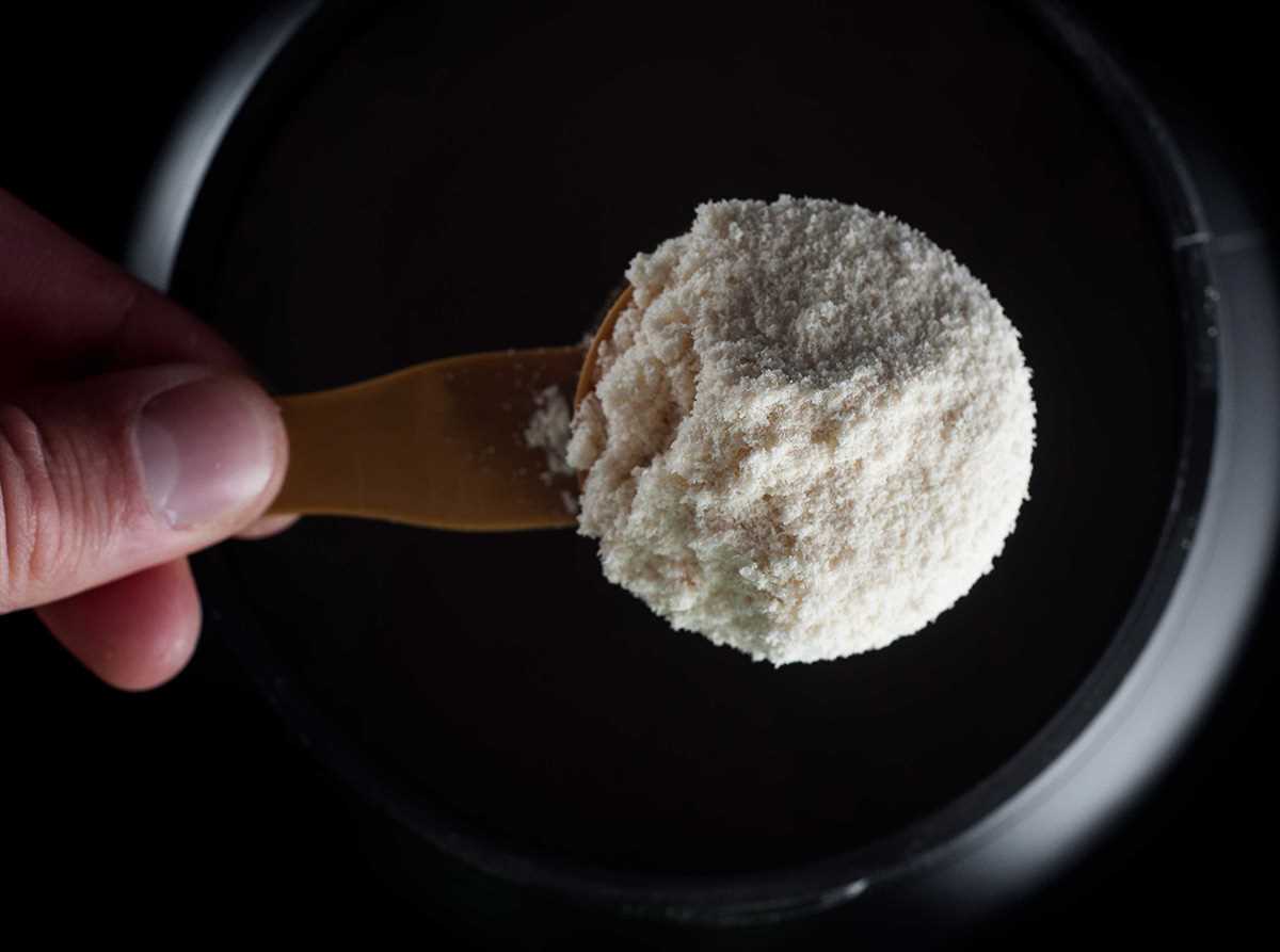 Pote de creatina em pó ao lado de uma colher medidora cheia, com o pó branco espalhado levemente sobre a mesa.