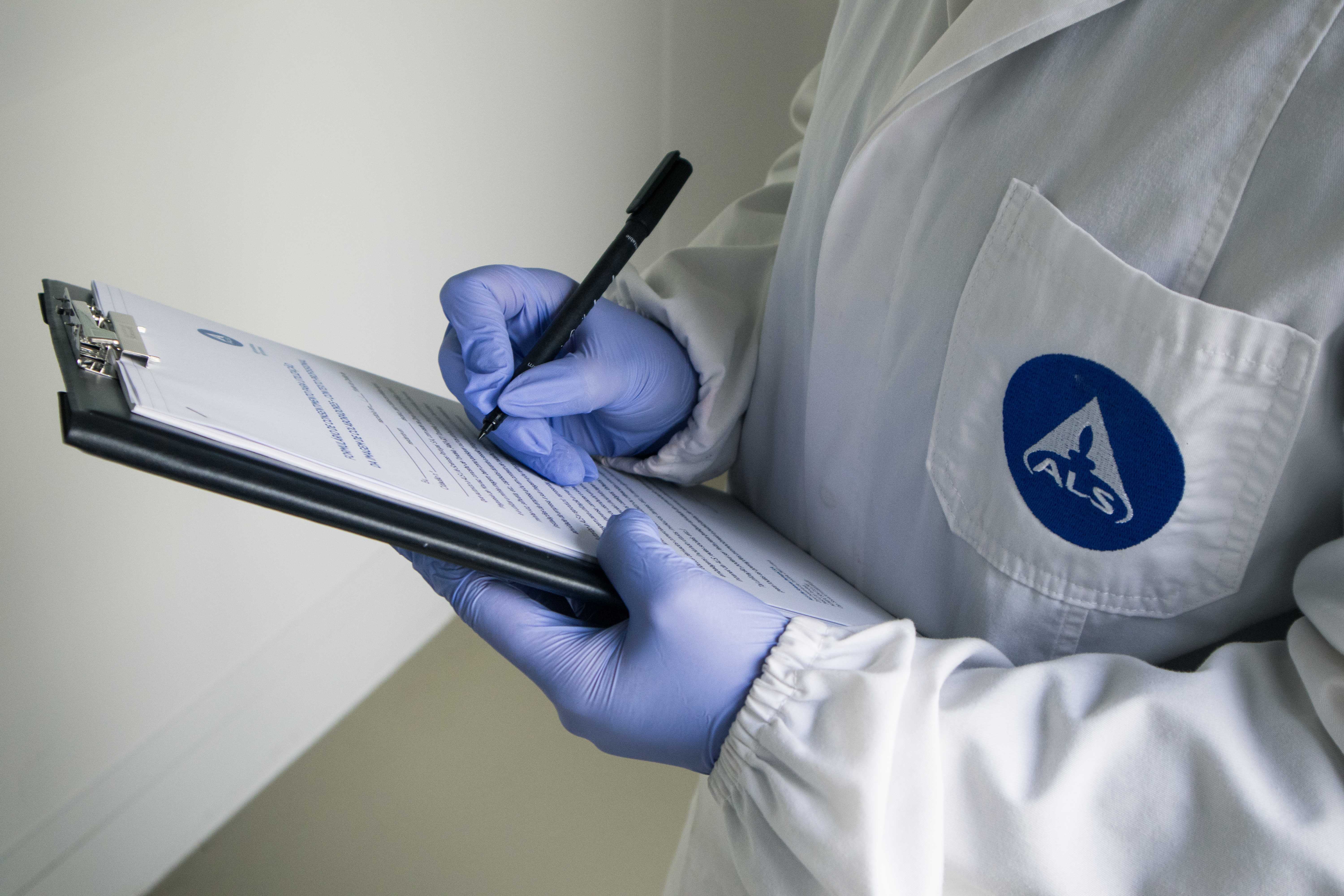 Técnico de laboratório com bata e luvas azuis a registar dados numa prancheta durante inspeção e controlo de qualidade em ambiente laboratorial.