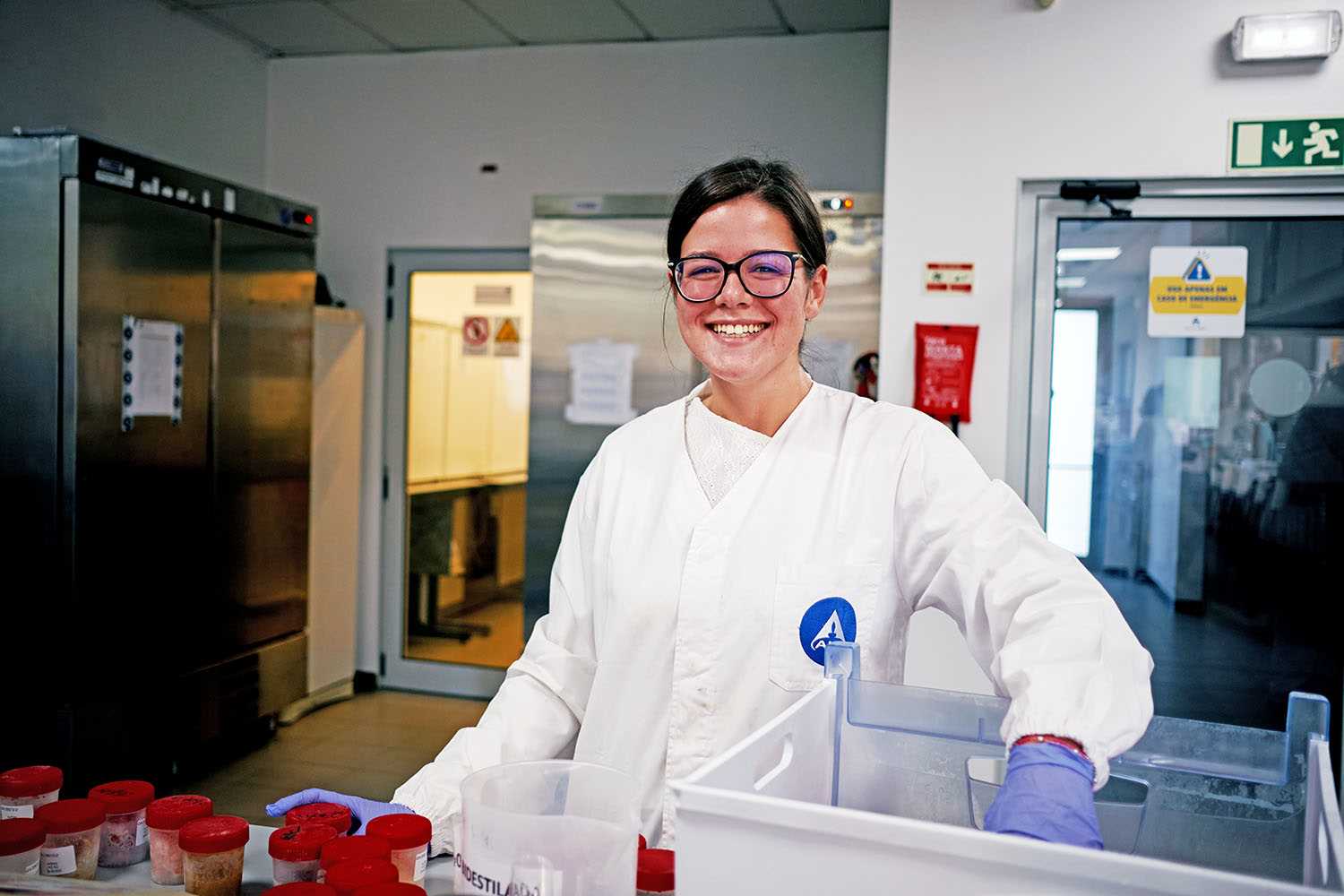 Pessoa com bata branca, luvas azuis e óculos em ambiente laboratorial, rodeada por equipamentos e recipientes, envolvida em atividades científicas ou de pesquisa.