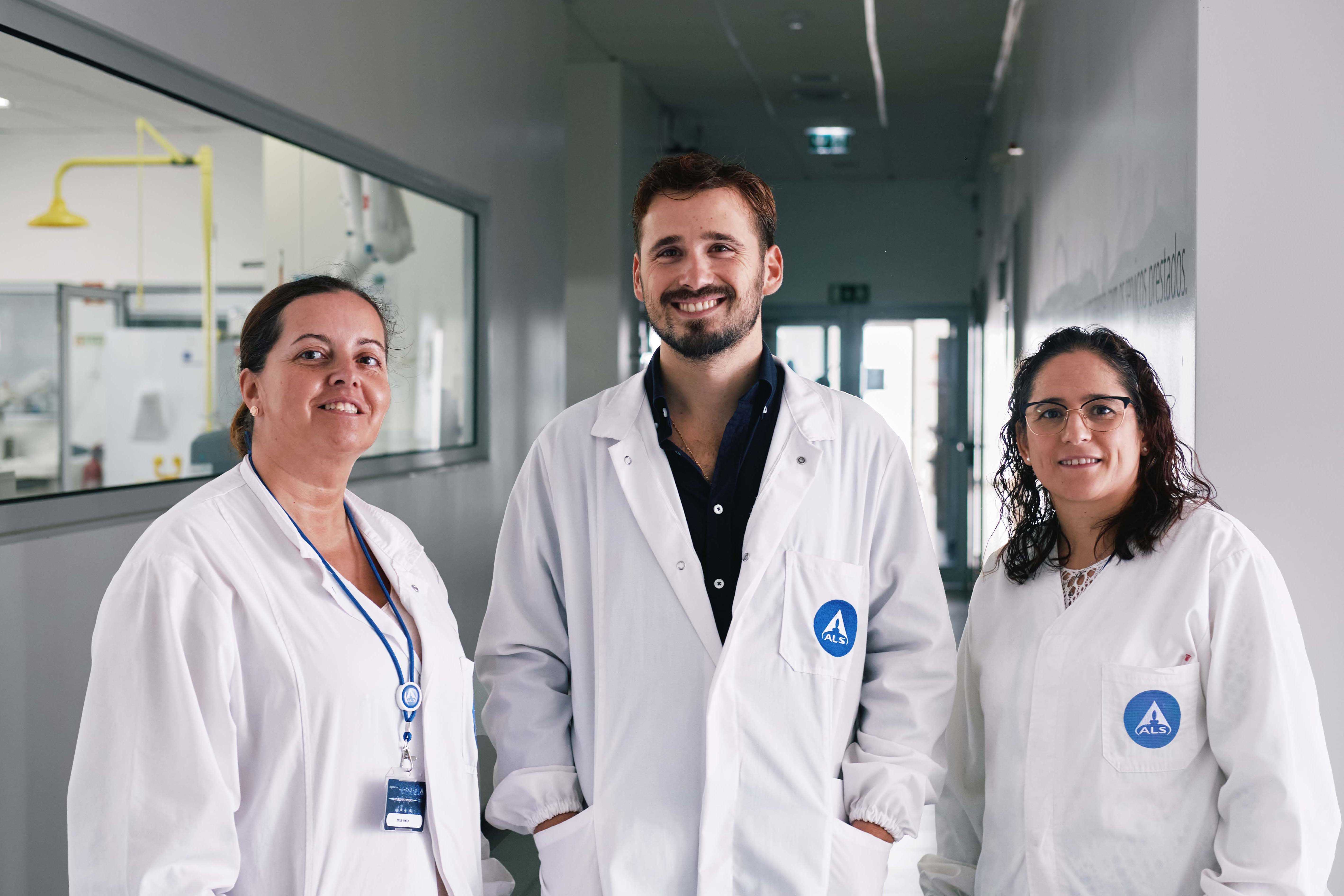 Três pessoas com batas brancas de laboratório a posar num corredor, com uma janela à esquerda mostrando parte de um espaço técnico.