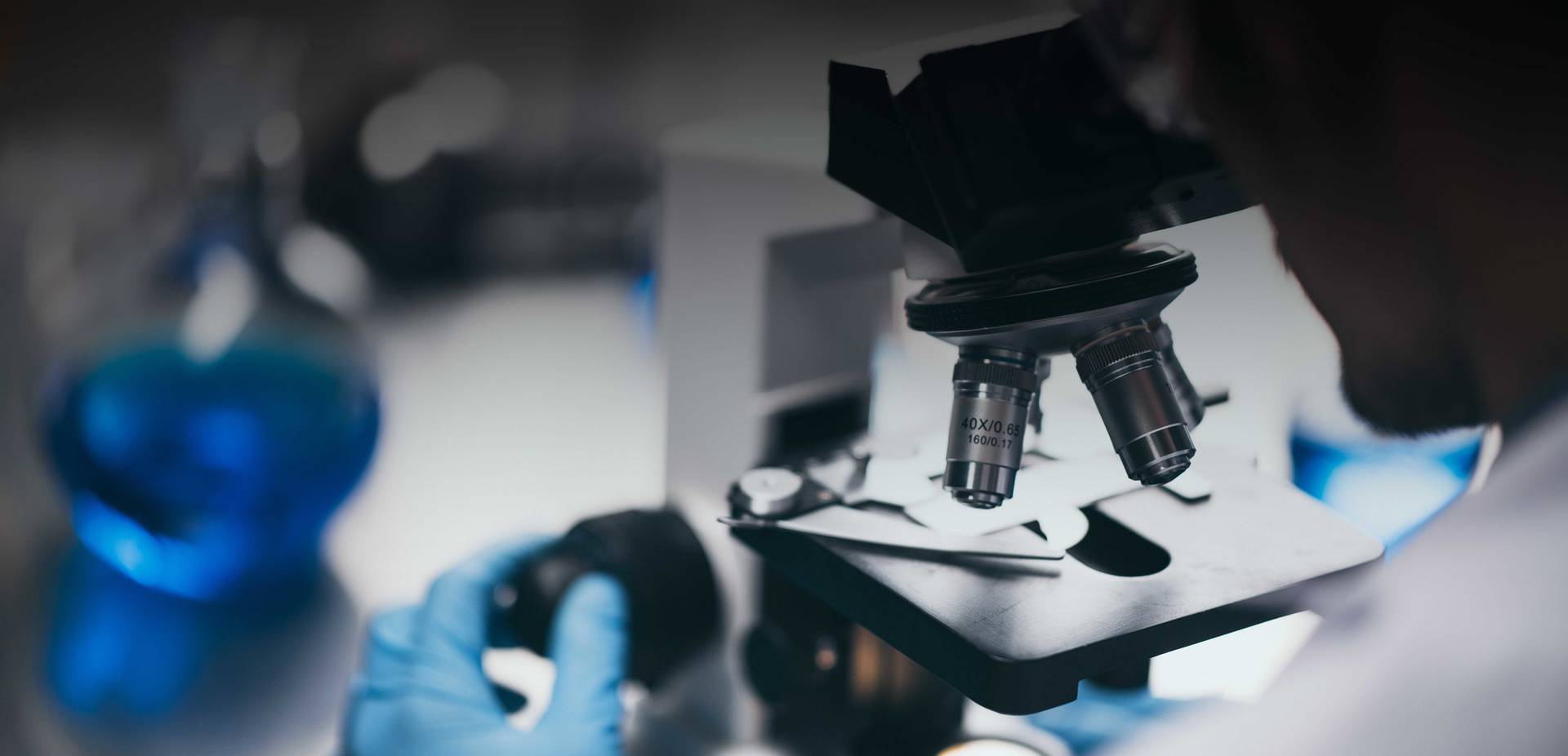 Laboratórny mikroskop v popredí s pripraveným preparátom na sklíčku; v pozadí je rozmazaná osoba v ochranných rukaviciach pracujúca v laboratórnom prostredí s modrými nádobami a vedeckým vybavením.