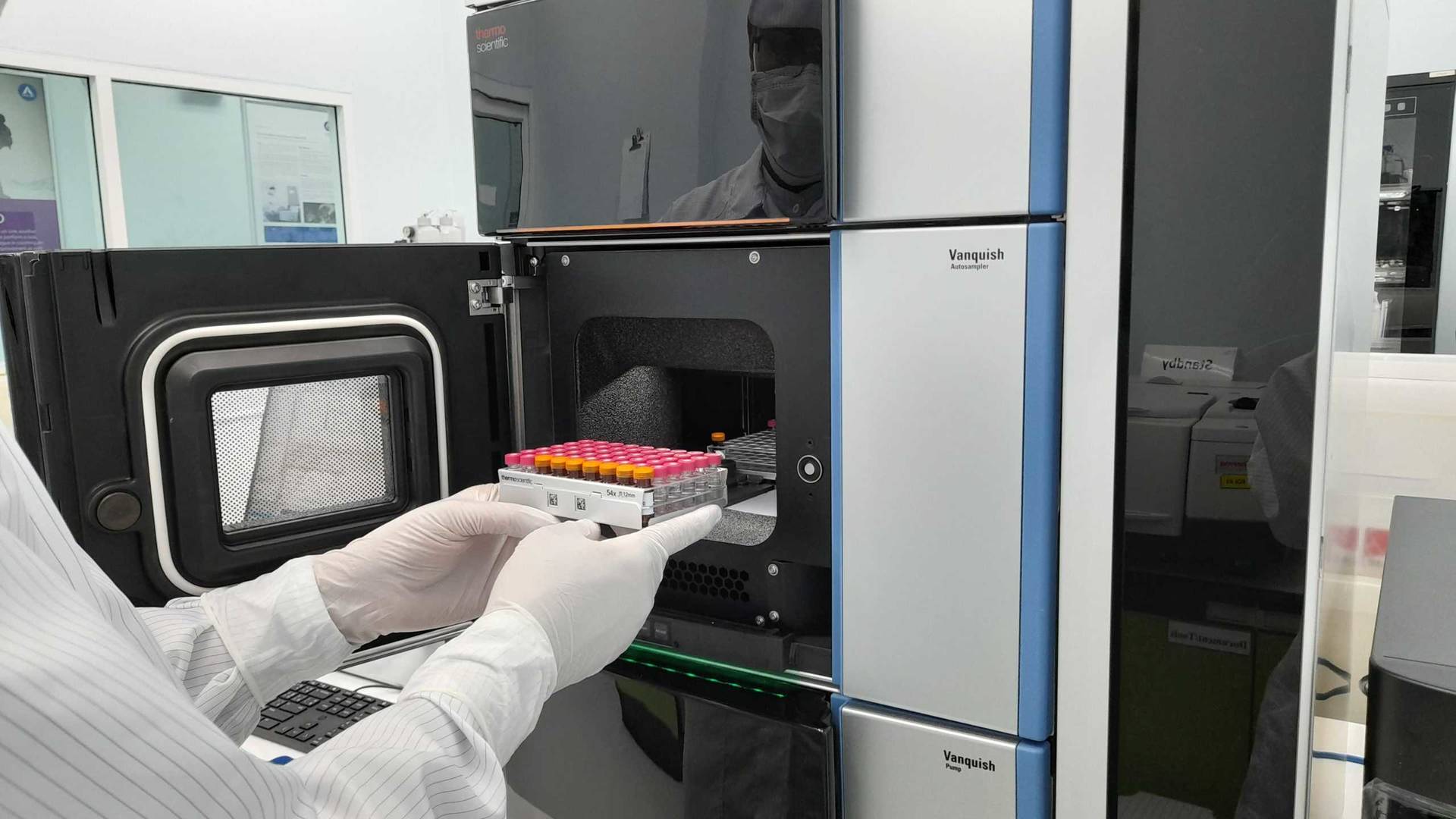 Gloved hands place a rack of sample vials into a high‑performance liquid chromatography (HPLC) system inside a clean laboratory environment.