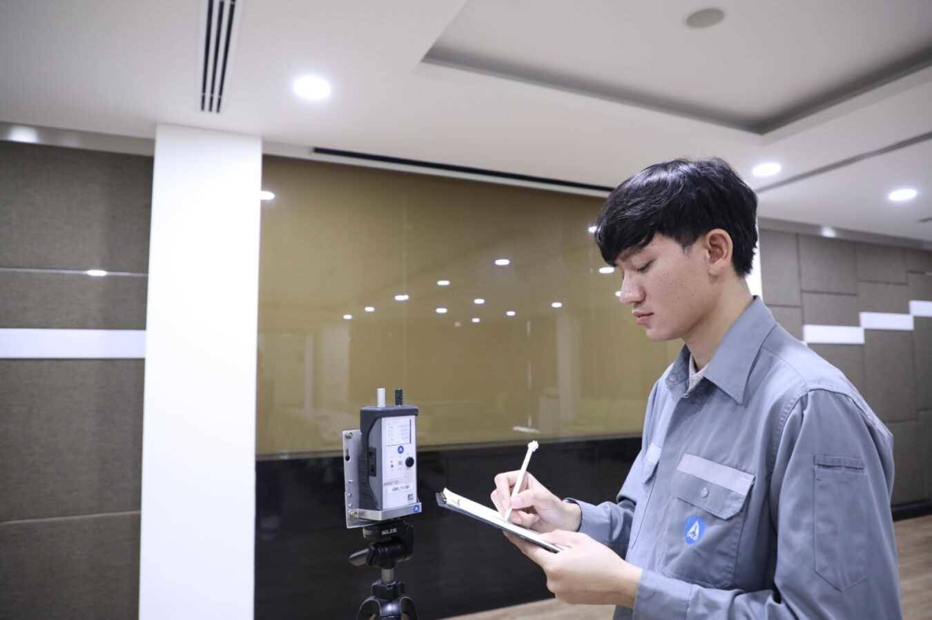 บุคคลสวมชุดทำงานกำลังบันทึกค่าการวัดลงบนแท็บเล็ตหรือคลิปบอร์ด ข้างอุปกรณ์ตรวจวัดคุณภาพอากาศภายในที่ติดตั้งบนขาตั้ง ภายในพื้นที่ทำงานภายในอาคาร