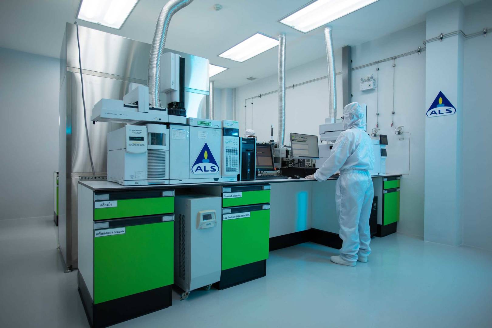 A person wearing a full cleanroom suit operates advanced laboratory analysis equipment in a bright, sterile testing laboratory, with green cabinets and ALS-branded instruments.