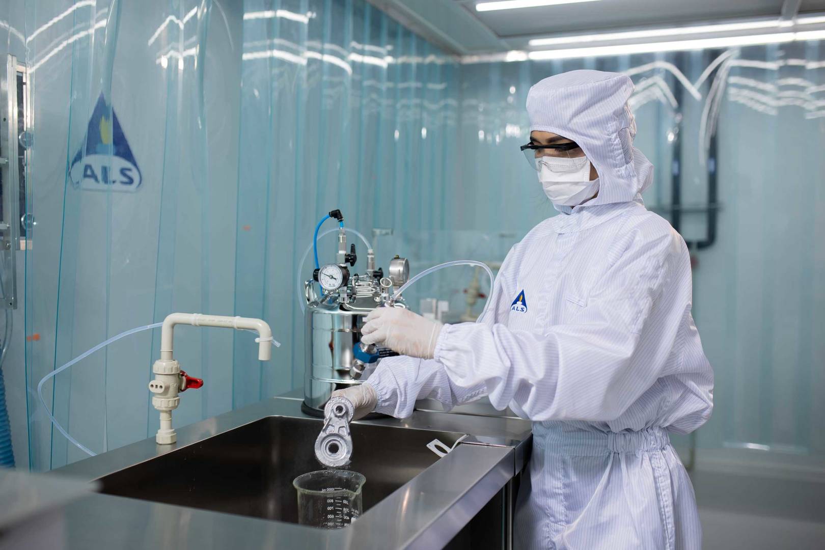 A person wearing cleanroom protective clothing operates a particle analysis system at a stainless‑steel sink, dispensing liquid into a beaker inside a controlled laboratory environment with ALS branding.