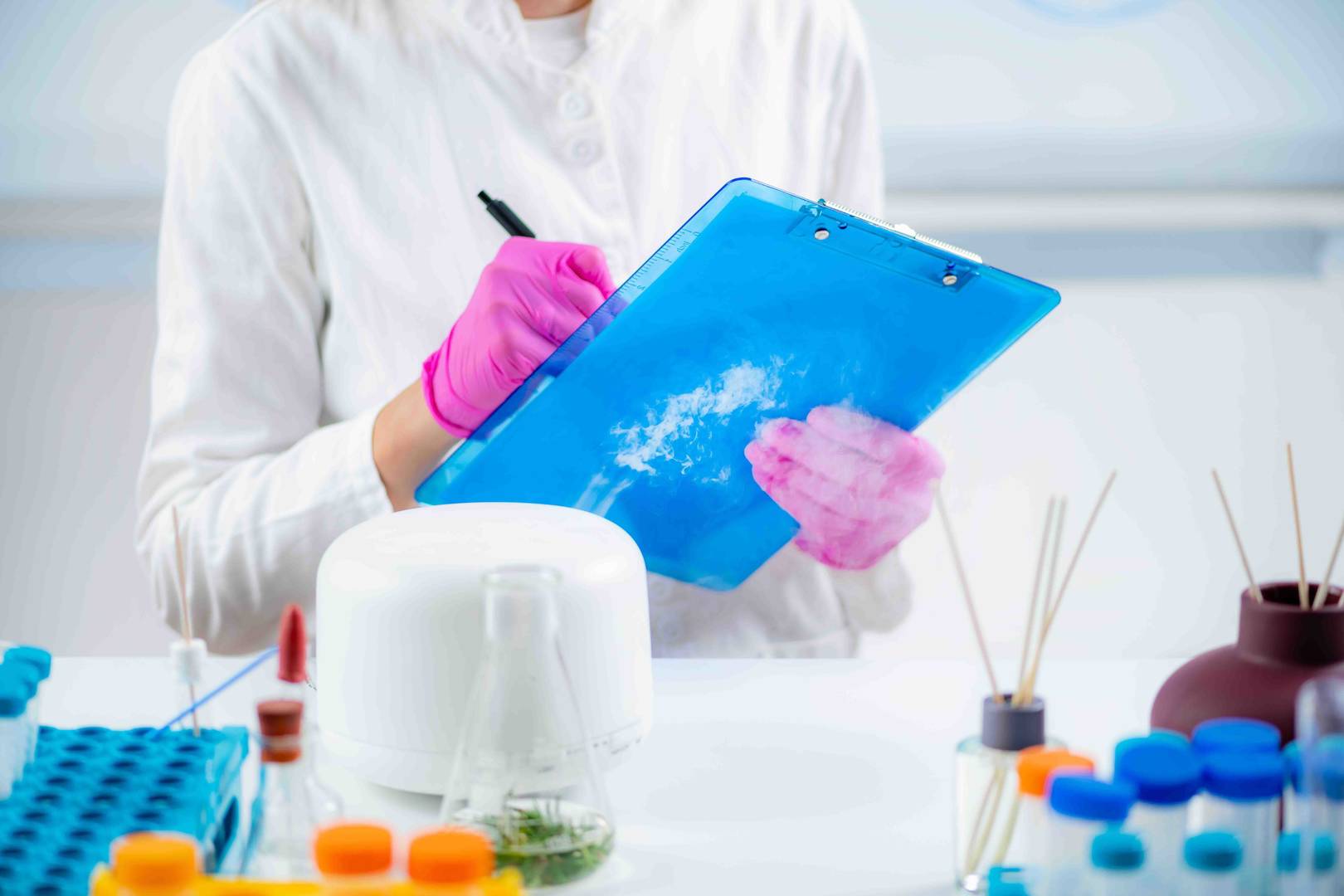 Laboratuvar ortamında mavi bir klipbord üzerine yazı yazan pembe eldivenli kişi.