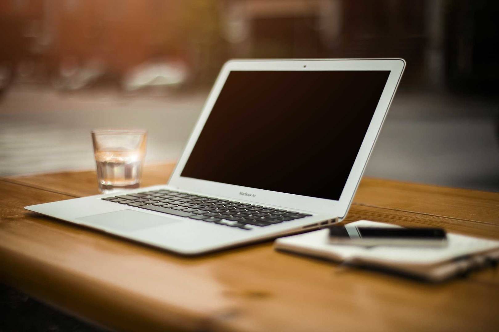 Laptop på brunt skrivbord. Ett glas med vatten på vänster sida och ett anteckningsblock med penna på höger sida om datorn