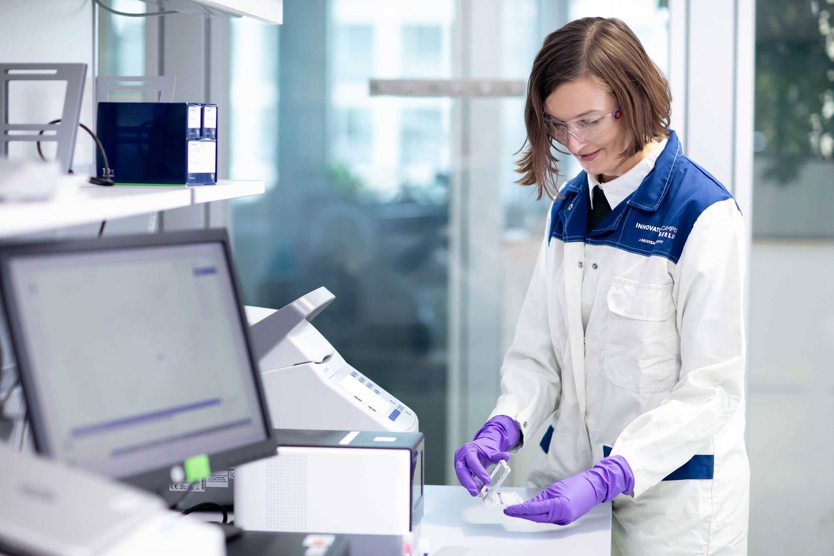 Laborantin steht im Labor vor einem Analysegerät und schaut sich eine Probe an. 